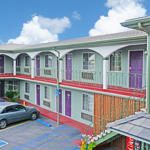 The image shows a two-story motel with parked cars, purple doors, balconies, red floors, and a blue sky with clouds in the background.