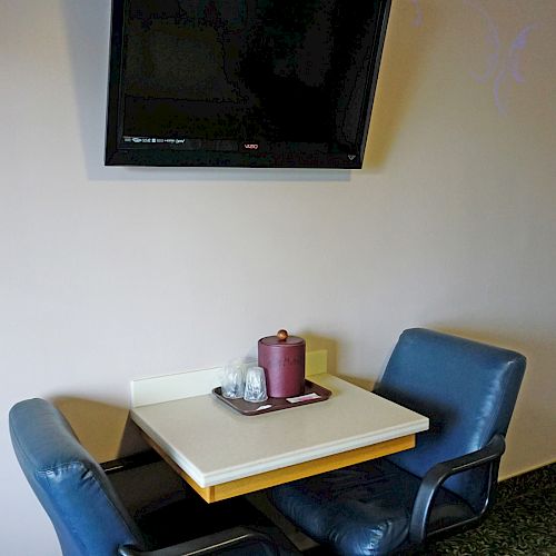 A small dining area with two blue chairs and a table, positioned below a mounted flat-screen TV on the wall. There are condiments on the table.