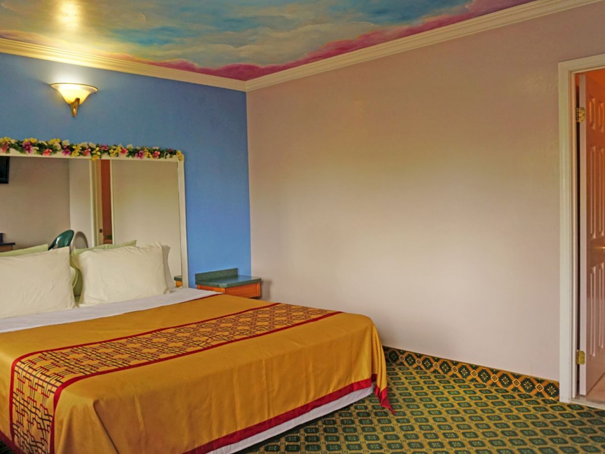 The image shows a hotel room with a large bed, blue accent wall, decorative ceiling, a mirror, a television, and an open door leading to a bathroom.