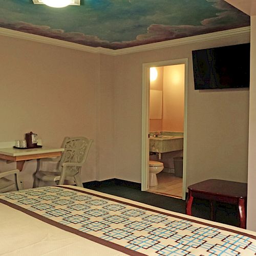 The image shows a hotel room with a bed, a small table with two chairs, a wall-mounted TV, and an open door leading to a bathroom.