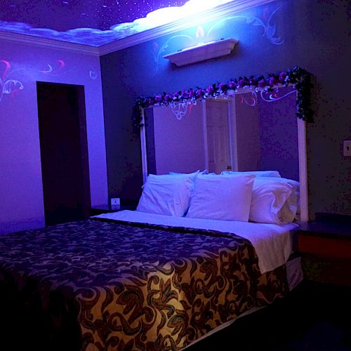 The image shows a dimly lit bedroom with a decorated bed, pillows, and a patterned blanket. The room has purple lighting and wall decorations.