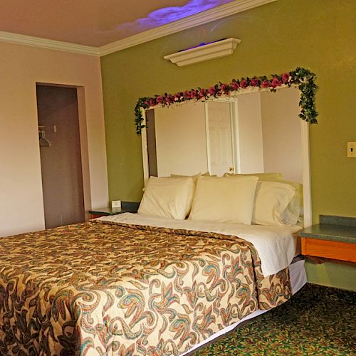 A hotel room featuring a decorated bed with a patterned cover, two pillows, a wall mirror with a floral garland, and a bedside table.