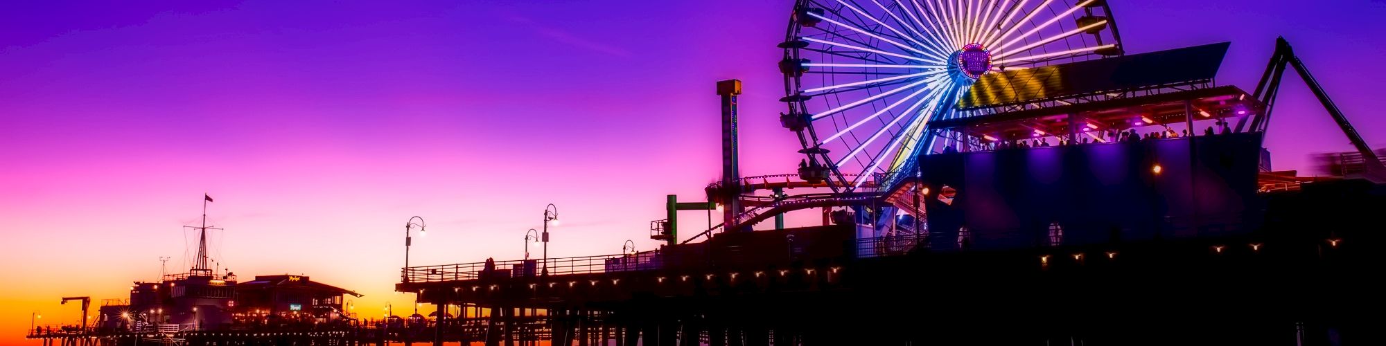 A vibrant sunset with a silhouetted pier, featuring a lit Ferris wheel and calm ocean waves in the foreground.