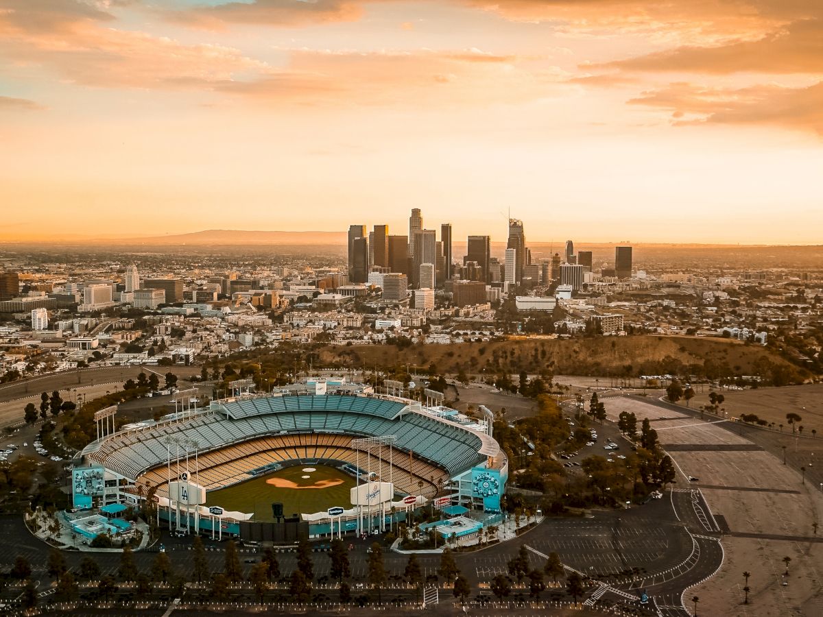 The image shows an aerial view of a large baseball stadium surrounded by parking areas, with a city skyline and a sunset in the background.