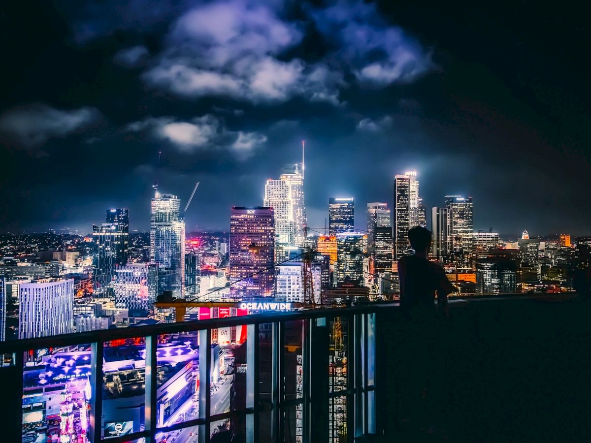 A person stands on a balcony overlooking a brightly lit city skyline at night, with tall buildings and neon lights under a partly cloudy sky.