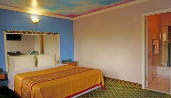 The image shows a hotel room with a double bed, a blue wall, ornate mirror, bathroom, and a scenic sky-painted ceiling.