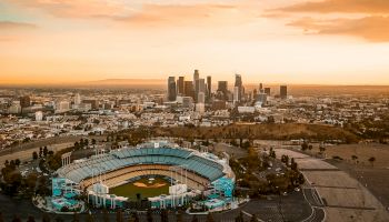 This image shows a baseball stadium in the foreground with a city skyline in the background under a vibrant sunset sky.