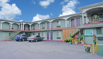 The image shows a two-story motel with parked cars, outdoor stairs, and potted plants decorating the entrance under a partly cloudy sky.