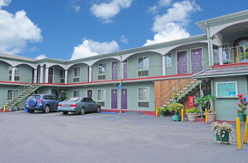 The image shows a two-story motel with parked cars, outdoor stairs, and potted plants decorating the entrance under a partly cloudy sky.