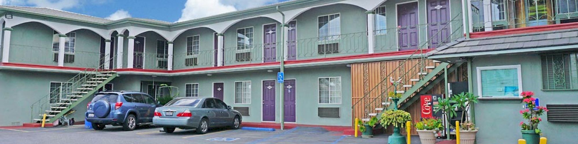 The image shows a two-story motel with parked cars, colorful doors, plants, and an outdoor stairway under a blue sky with clouds.