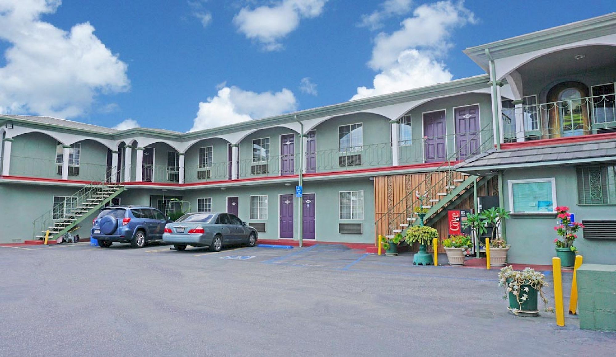 A motel with pastel-colored rooms, exterior staircases, a parking lot, blue sky, and potted plants along the walkway, two cars parked.