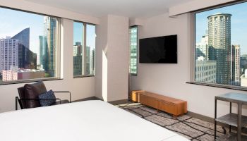 The room features large windows with city views, a wall-mounted TV, a bed, a chair, and a wooden bench.
