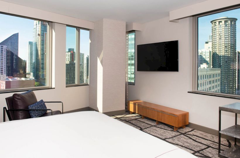 The room features large windows with city views, a wall-mounted TV, a bed, a chair, and a wooden bench.