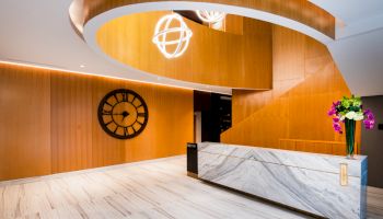 The image shows a modern lobby with wood paneling, a large clock on the wall, a curved staircase, and a marble reception desk with flowers.