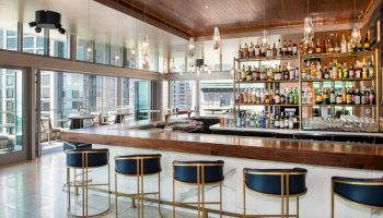 This image shows a modern bar with high stools, a wooden counter, and a well-stocked shelf with various bottles of alcohol, in a bright setting.