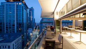 A modern balcony attached to a high-rise building, overlooking a cityscape with tall buildings and a street below at dusk.