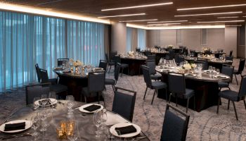 A banquet hall with round tables set with black napkins, white flowers, glassware, and silverware under modern lighting.