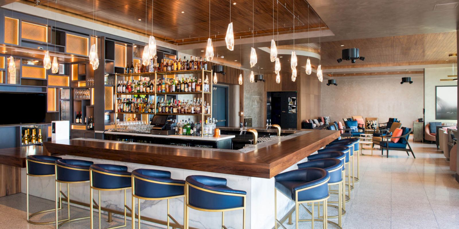 A modern bar with a well-stocked counter, stylish bar stools, hanging lights, and seating area in a contemporary, bright setting.