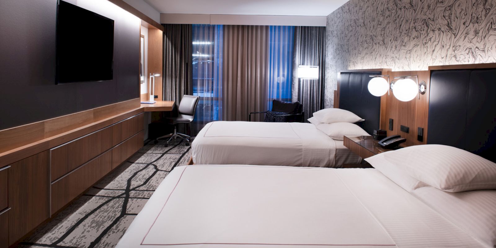 The image shows a modern hotel room with two double beds, a TV, desk with a chair, and stylish decor featuring patterned wallpaper and ambient lighting.