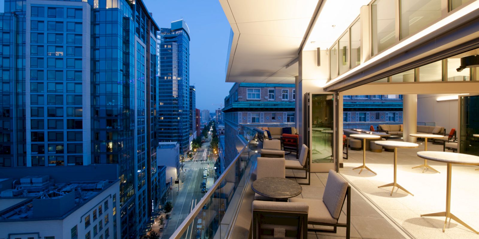 A modern balcony with seating and tables overlooks a busy city street lined with tall buildings at dusk, with lights illuminating the urban scene.