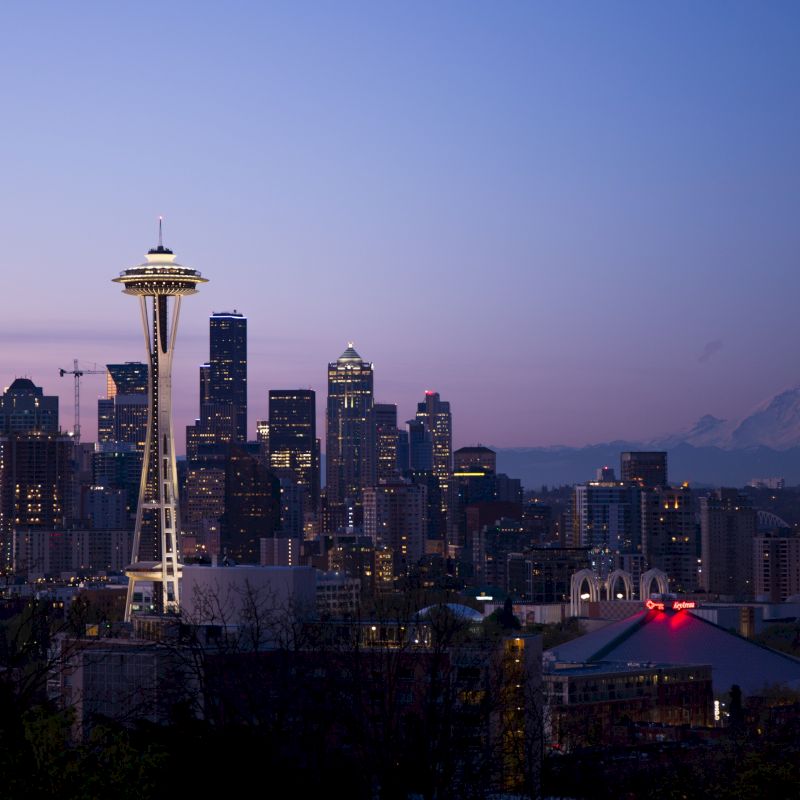 A city skyline at dusk with illuminated buildings, the Space Needle prominently visible, and a distant mountain against a purple sky in the background.