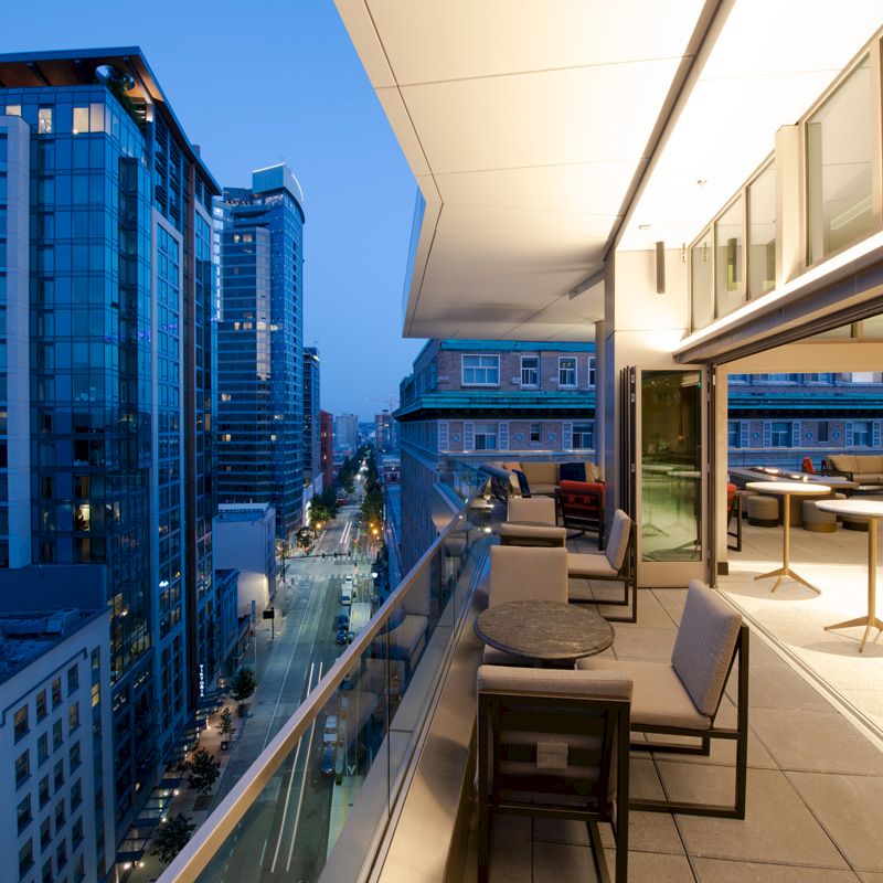 A modern cityscape at dusk is shown from a balcony with outdoor seating and tables, overlooking a busy street and surrounding high-rise buildings.