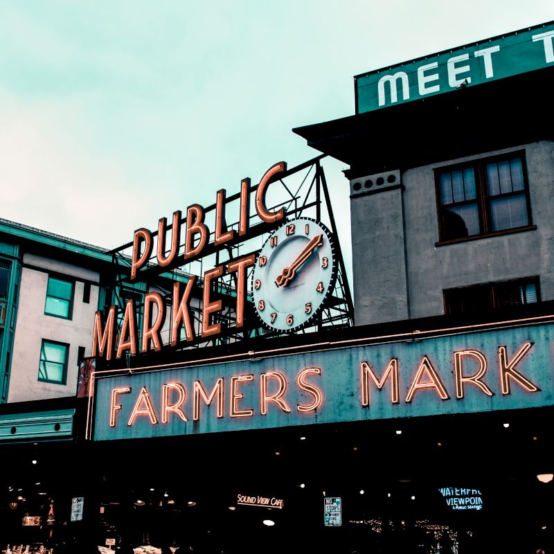 The image shows the iconic sign of the Public Market and Farmers Market at Pike Place Market in Seattle, Washington, with older buildings in the background.
