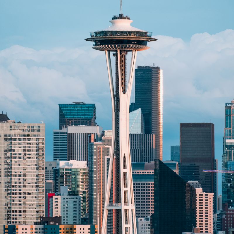 The image shows the iconic Space Needle in Seattle, surrounded by various buildings and skyscrapers against a backdrop of clouds and blue sky.