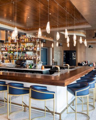 This image shows a modern bar with a wooden counter, blue cushioned stools, hanging lights, and a variety of bottles on display.