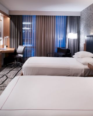 The image shows a modern hotel room with two double beds, a desk near the window, a flat-screen TV mounted on the wall, and contemporary decor.