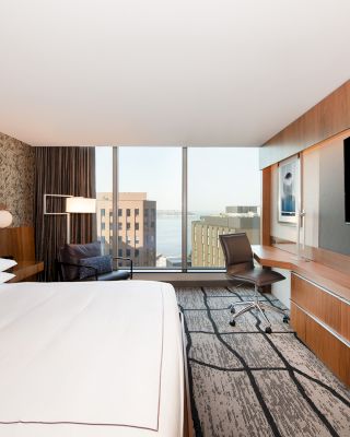 A modern hotel room with a large bed, patterned wall, desk and chair, flat-screen TV, and a window showing a cityscape view.