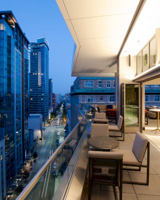 The image shows a modern rooftop terrace with outdoor seating and tables, overlooking a cityscape of tall buildings during twilight hours.