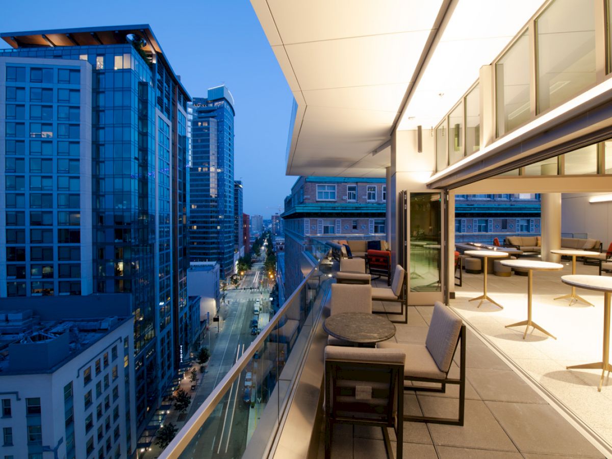 A modern cityscape at dusk, viewed from a balcony with seating and tables. The buildings are illuminated and the sky is a deep blue.