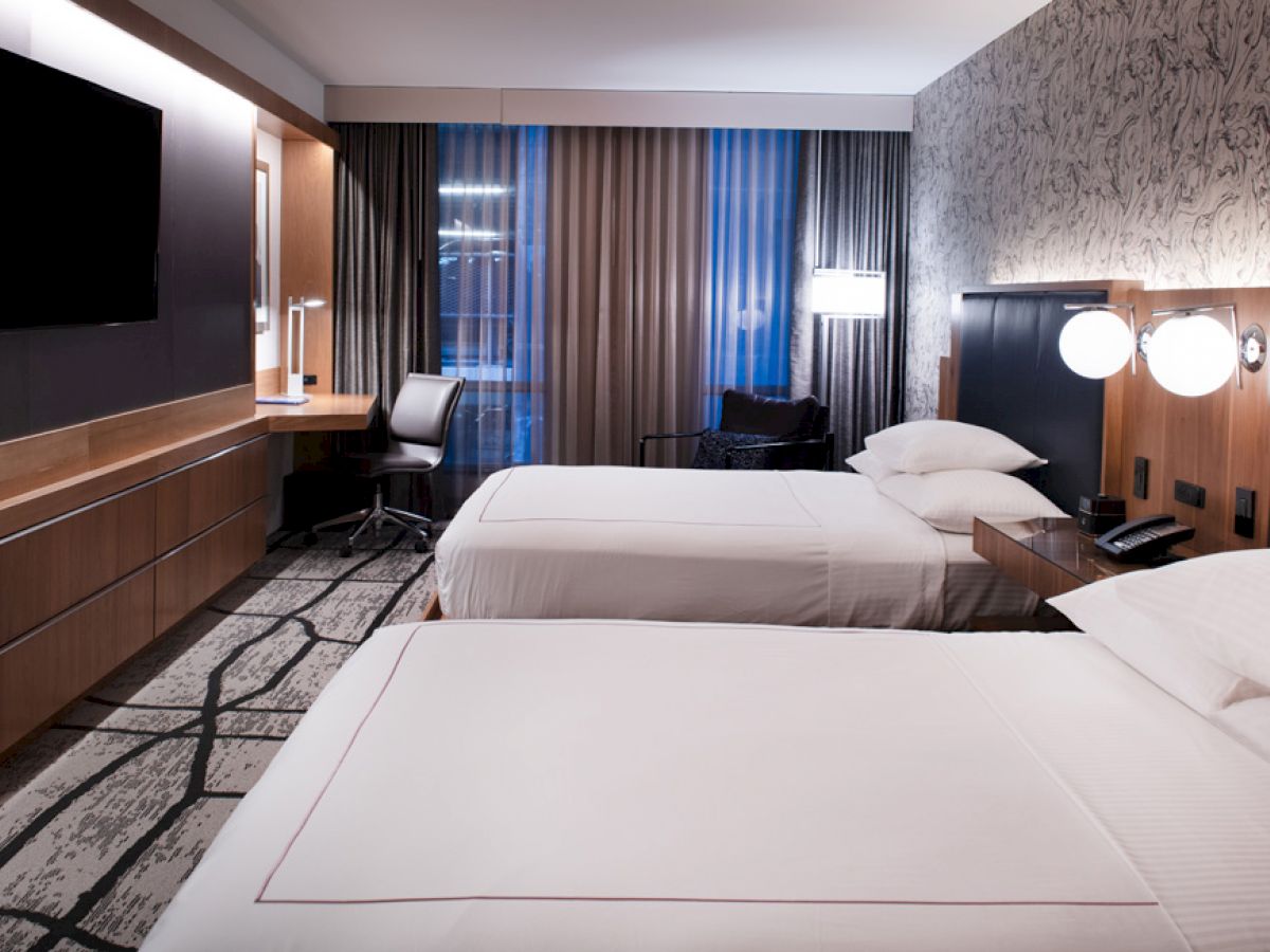 A modern hotel room featuring two neatly made beds, a large TV, a desk with a chair, and contemporary decor with subtle lighting.