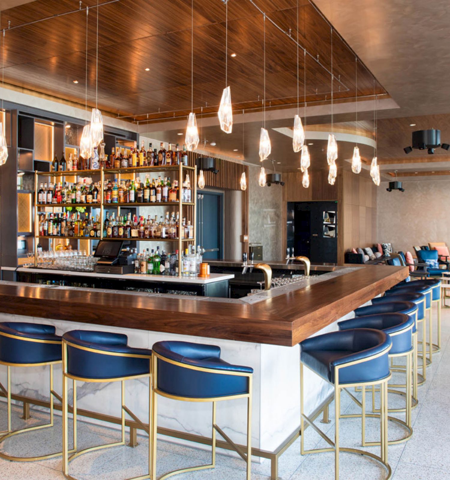 The image shows a modern bar with a wooden counter, blue chairs, hanging lights, and a well-stocked back bar. The room has a sleek, contemporary design.