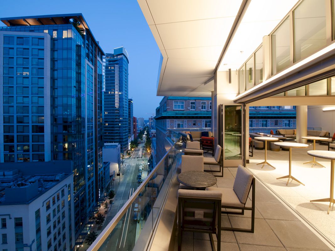 A modern urban balcony with outdoor seating overlooks a city street lined with tall buildings during twilight, featuring a sleek design and lighting.
