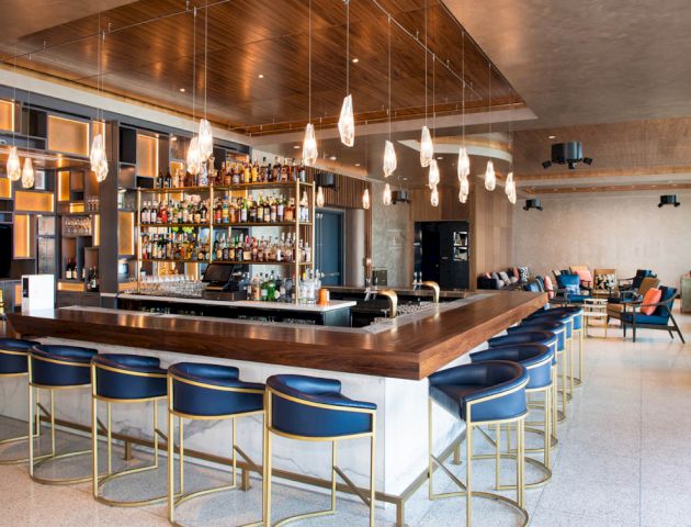 A modern bar with a wooden countertop, blue stools, hanging lights, and a fully stocked back bar, surrounded by a stylish seating area.