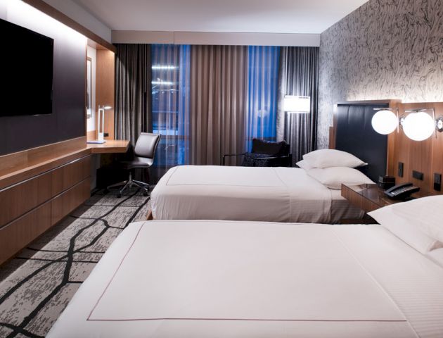 A modern hotel room with two beds, a wall-mounted TV, a desk, chair, and stylish lighting and decor.