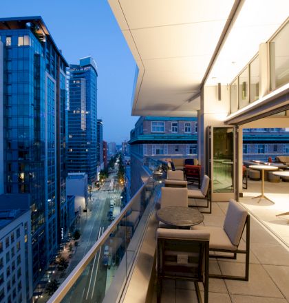 This image shows a modern balcony with seating arrangements overlooking a cityscape with tall buildings and a street below at dusk.