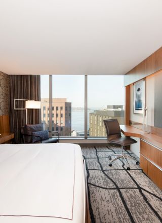 A modern hotel room with a large bed, desk, chair, flat-screen TV, and a city view through floor-to-ceiling windows ending the sentence.