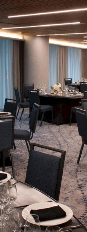 The image shows an elegantly set banquet hall with round tables, black chairs, white flower centerpieces, neatly arranged tableware, and ambient lighting.