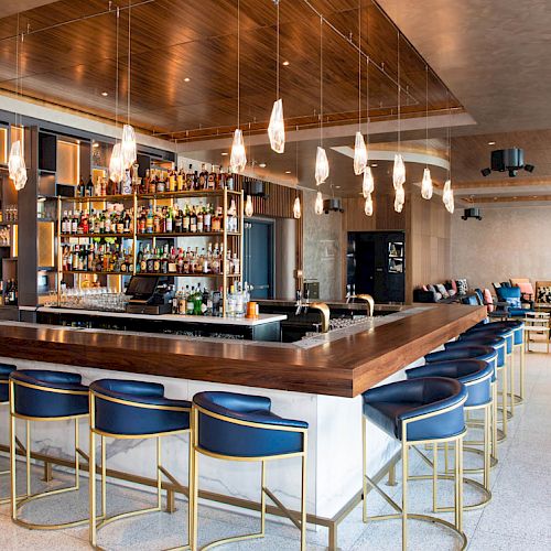 The image shows a modern bar with a wooden counter, blue barstools, and various hanging lights. Bottles are stocked on shelves behind the counter.