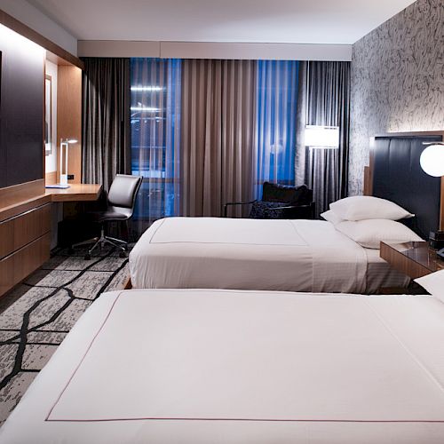 A modern hotel room with two double beds, a wall-mounted TV, a desk, and a stylish design featuring patterned wallpaper and contemporary furniture.