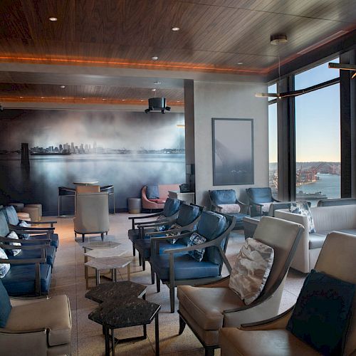 The image shows a modern lounge area with leather seating, large windows offering a water view, and a mural of a city skyline on one wall.