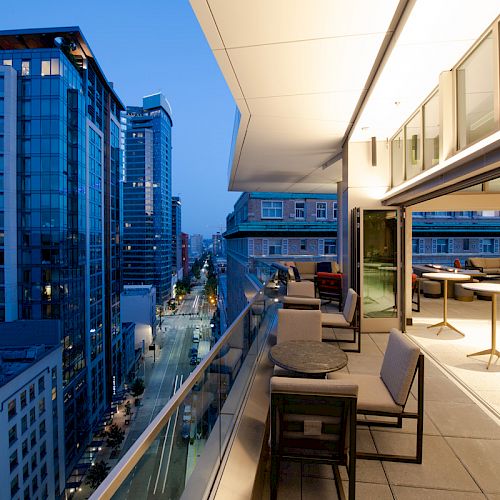 A modern balcony with seating areas overlooks a cityscape with tall buildings and a street below under dusk sky.