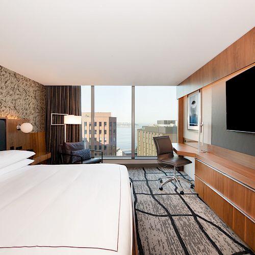 A modern hotel room with a large bed, patterned wall, desk, chair, flat-screen TV, and a window with a cityscape view outside.