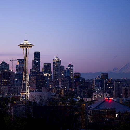 A twilight cityscape with the Space Needle in Seattle, featuring illuminated buildings and Mount Rainier in the background against a purple sky.