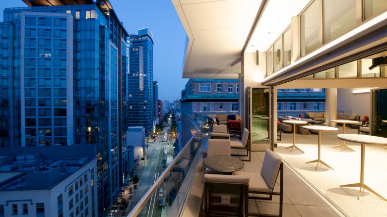 An evening cityscape viewed from a modern balcony with tables, chairs, and buildings illuminated. The street and skyscrapers are visible below.