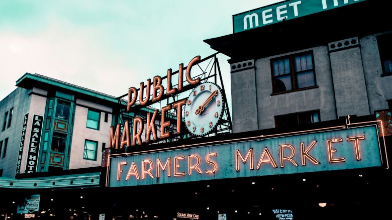The image shows a public market with neon signs for "Public Market" and "Farmers Market" with a clock. A building in the background reads "MEET THE.".