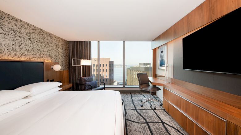 A modern hotel room with a large bed, a desk with a chair, a flat-screen TV, and a city view through the large window.